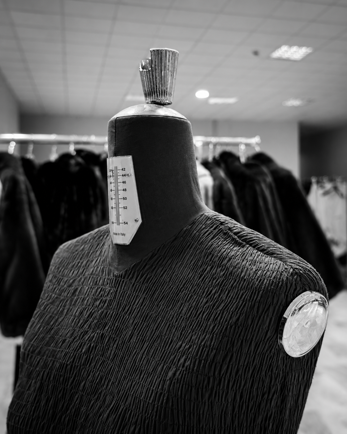 A tailor’s dress form marked with a sizing scale, standing in front of rails of finished fur coats in the Casiani workshop in Kastoria